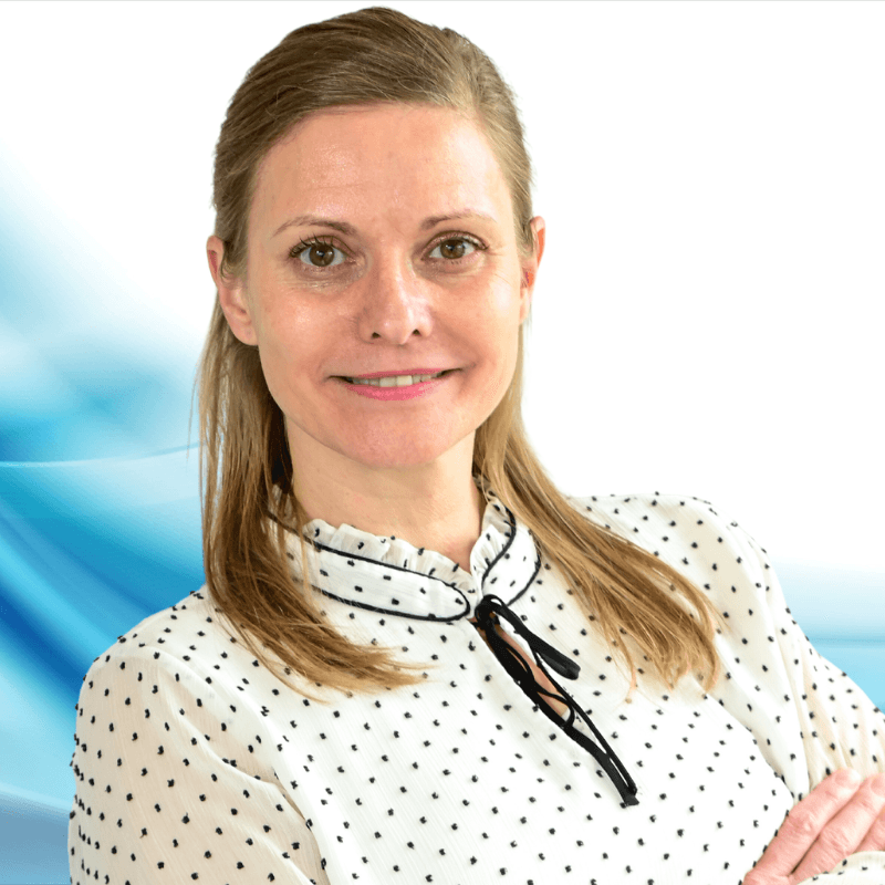 Smiling woman with light brown hair in a white polka-dot blouse, arms crossed against a blue abstract backdrop.