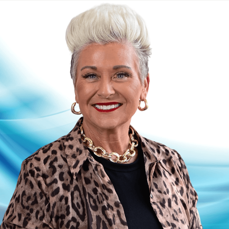 Smiling mature woman with short platinum hair wearing a leopard print jacket and gold necklace, portrait against an abstract blue background.