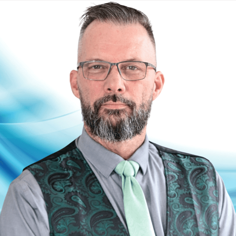 Professional headshot: bearded man with glasses, gray shirt, green tie, and a green paisley vest against a blue abstract background.